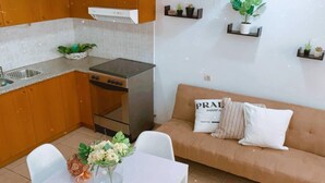 Eine kompakte Küche mit Herd, Backofen und Spüle. Ein Esstisch mit Stühlen und eine Blumenvase stehen bereit. Gegen die Wand lehnt eine Couch mit Kissen.