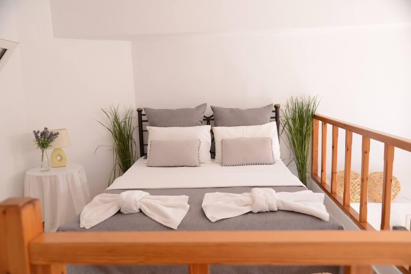 A bed with a wooden frame, grey bedding, and two pillows. There is a small round table with a lamp and a vase of flowers to the left.