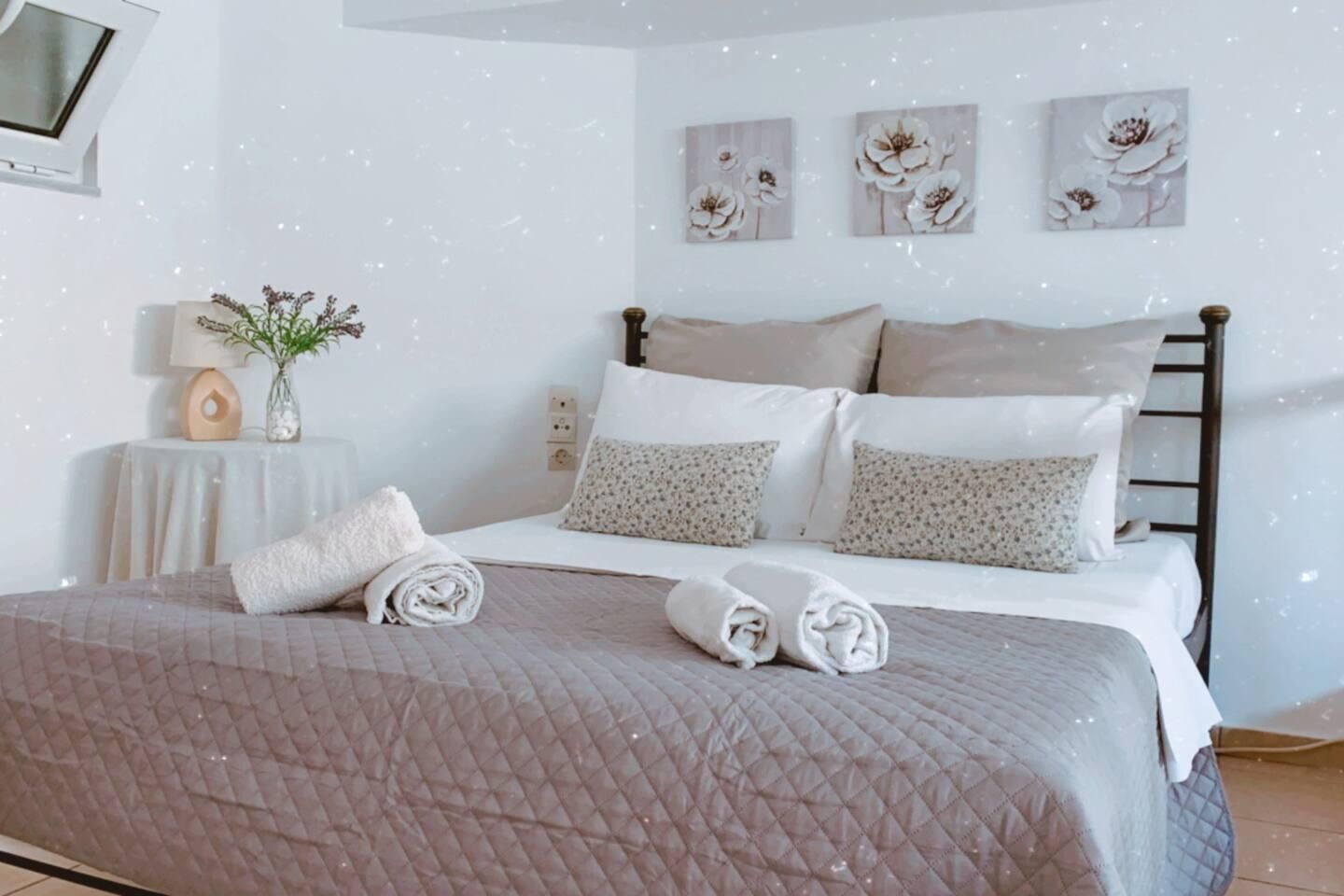 Un lit bien fait, avec des serviettes pliées, une table de chevet avec une lampe et un vase, et trois œuvres d’art florales accrochées au mur.