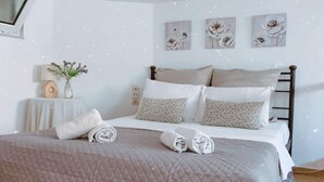 Ein ordentlich bezogenes Bett mit aufgerollten Handtüchern, ein Nachttisch mit Lampe und Blumenvase sowie drei Blumenbilder an der Wand.