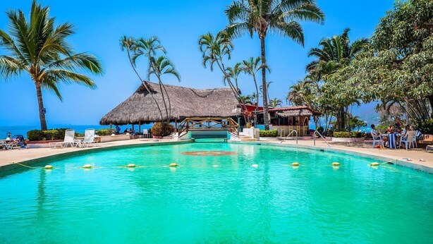 Une piscine à l’eau turquoise et limpide, entourée de palmiers et bordée d’une structure à toit de chaume.