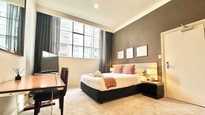 A hotel room with a bed, bedside table, lamp, and a desk with a chair. - Perfect City Pad (Auckland)
