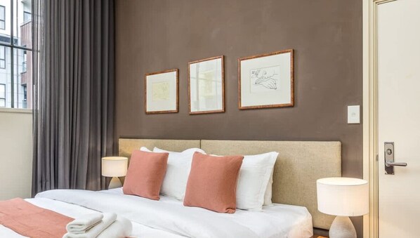A hotel room with a bed, two pillows, a nightstand, a lamp, and three framed artworks on the wall. - Perfect City Pad (Auckland)