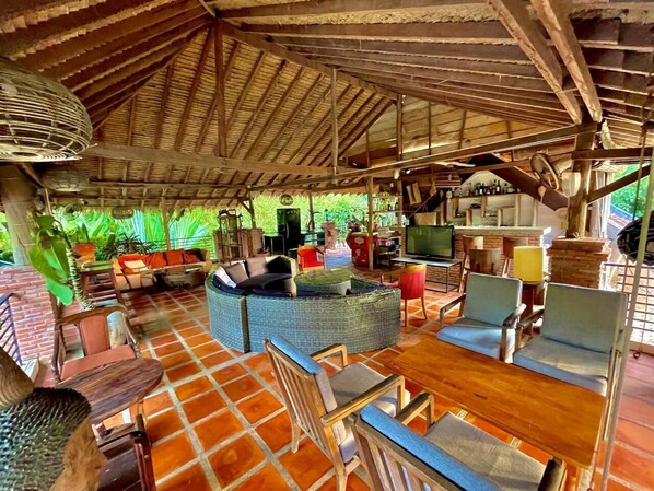 A covered outdoor lounge area with a thatched roof, wicker furniture, and a brick floor.