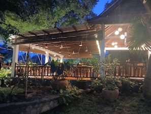 Un área de patio techada al aire libre con piso de madera, plantas en macetas y lámparas colgantes.