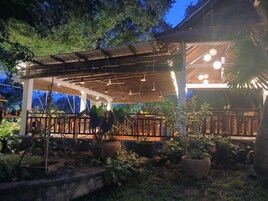 Un área de patio techada al aire libre con piso de madera, plantas en macetas y lámparas colgantes.