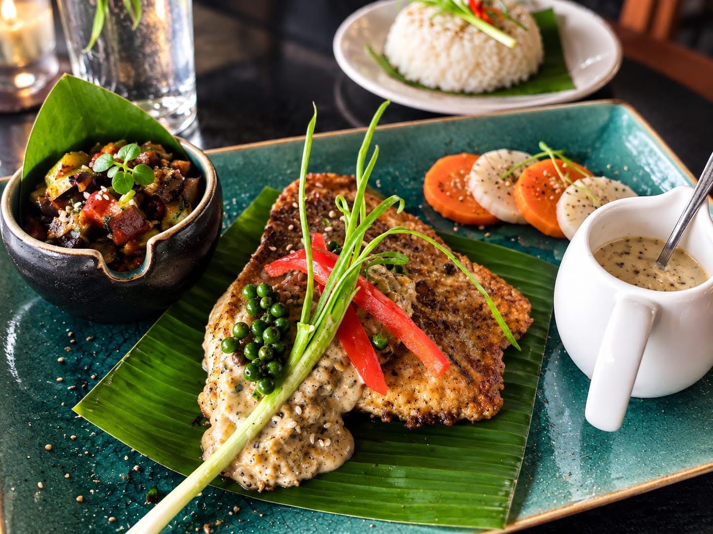 A meal consisting of a grilled fish fillet with sauce, a bowl of vegetable curry, sliced tomatoes, and a cup of sauce on a green leaf.