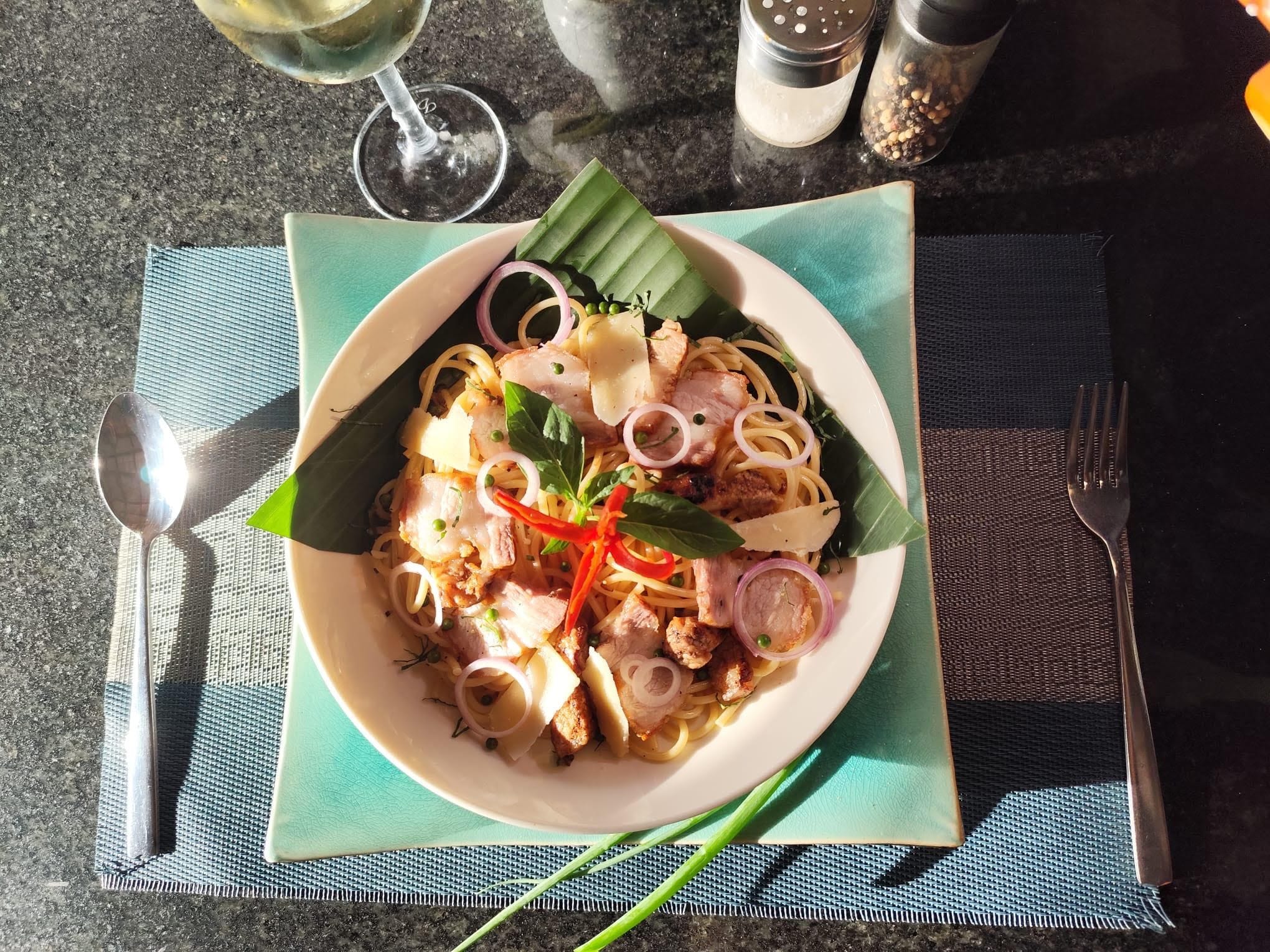 Un bol de nouilles avec de la viande et des légumes, une cuillère, une fourchette et un verre de vin blanc sur une table.