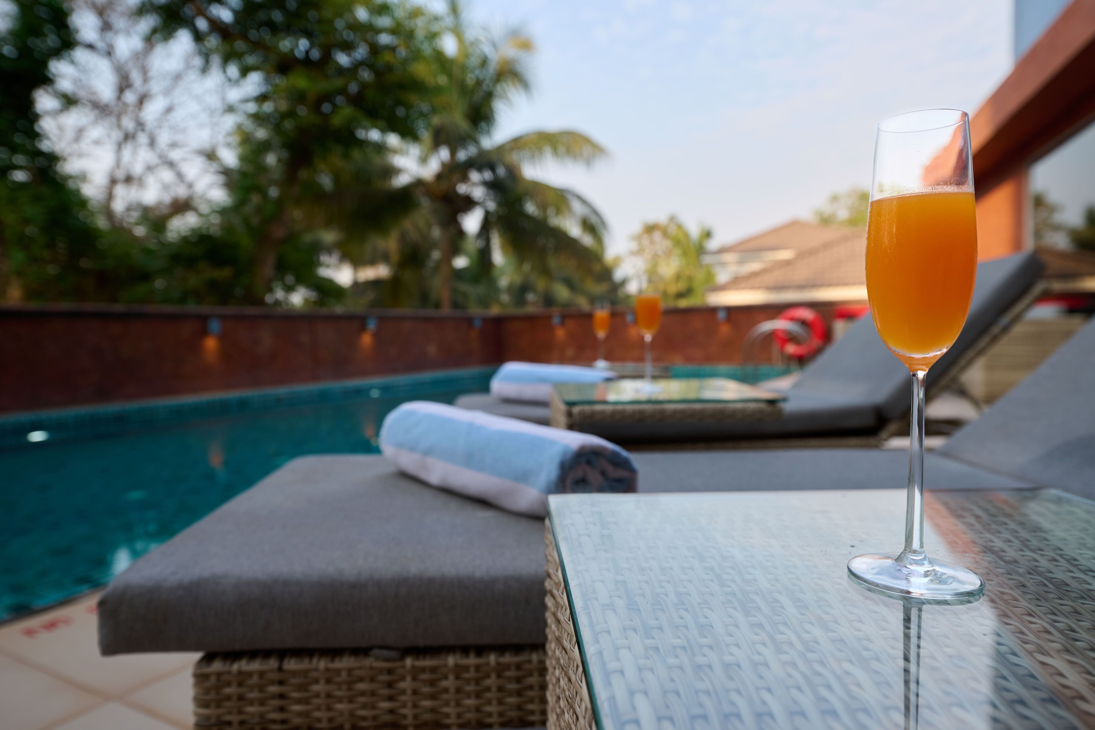 A glass of orange juice on a glass table near a poolside lounge area with cushions and a wicker chair.