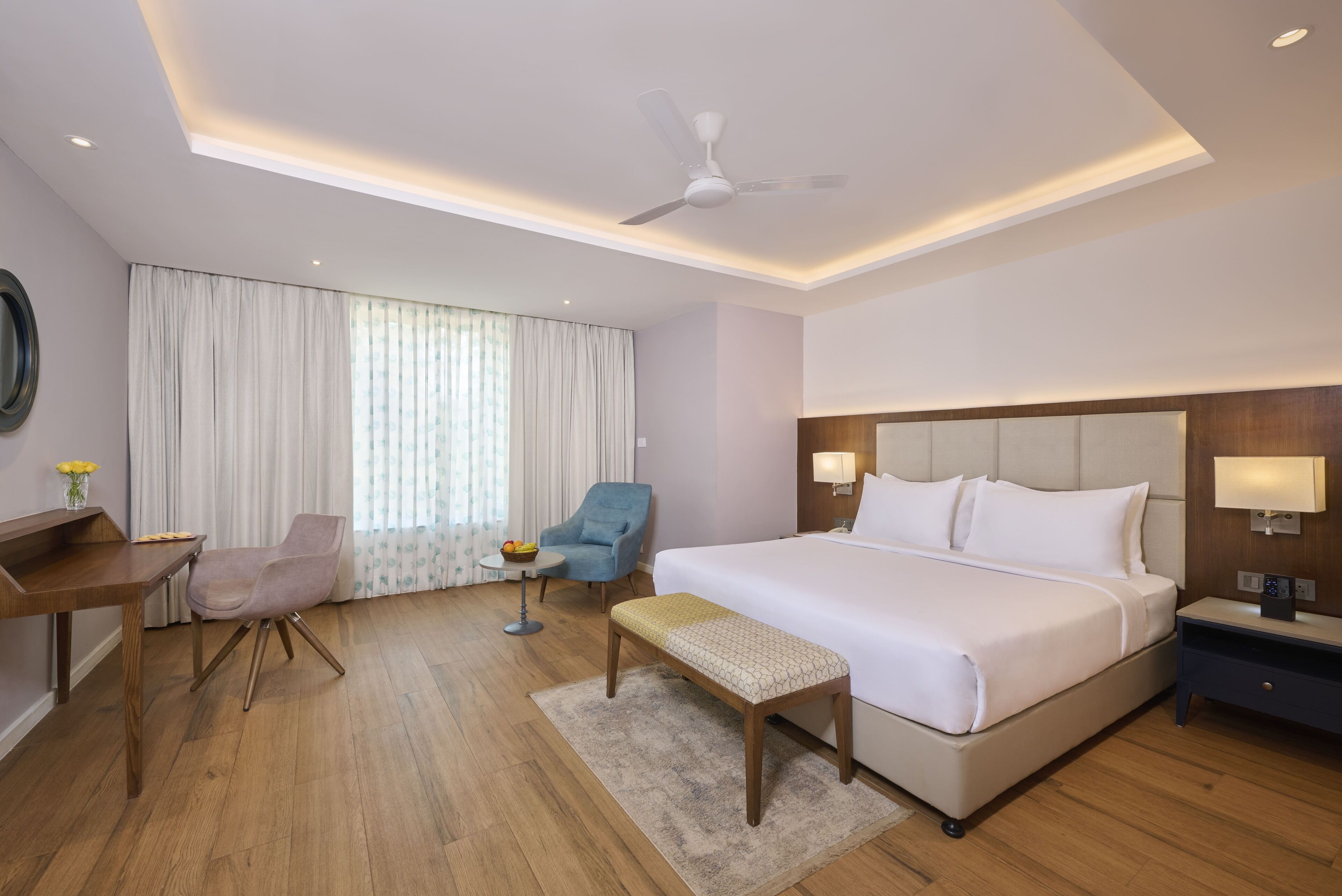 Habitación de hotel con una cama grande, un escritorio, una silla, una mesita con un frutero, un banco y un ventilador de techo.