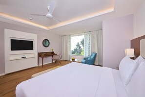 A hotel room with a large bed, a desk with a TV, a chair, a small table with fruit, and a window with curtains.