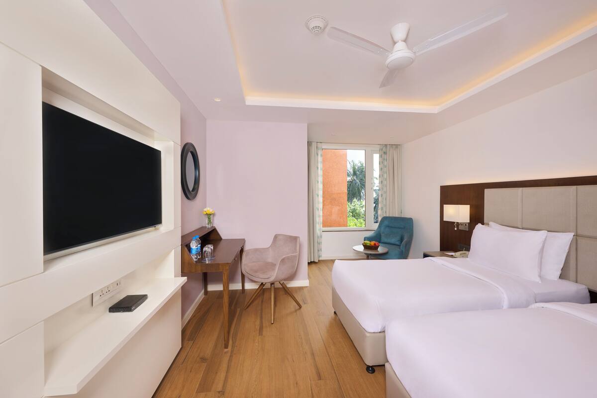 A hotel room with a bed, a desk, a chair, a TV, and a window with a view of greenery.