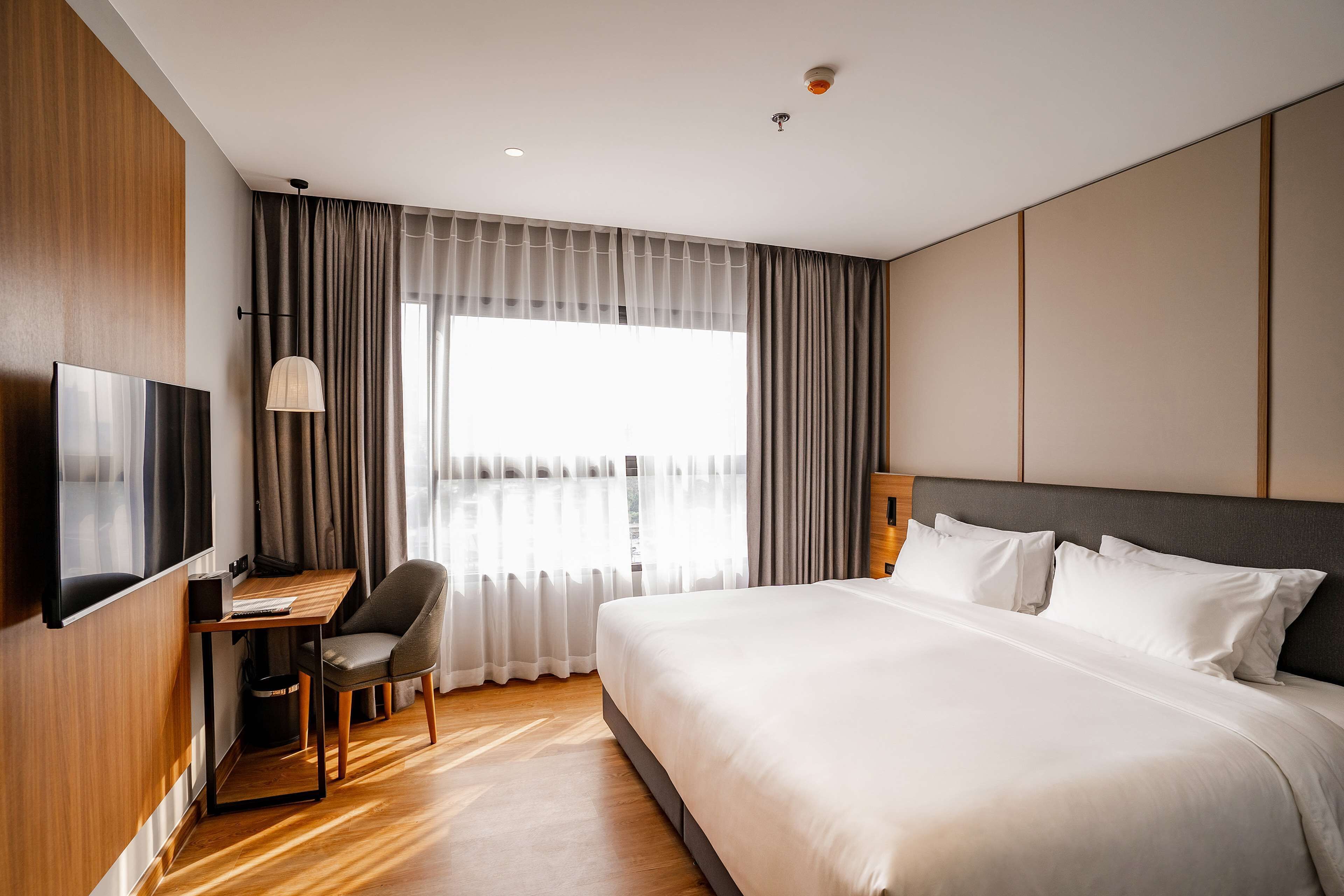 A modern hotel room with a large bed, a desk with a chair, a TV, and a large window with curtains.