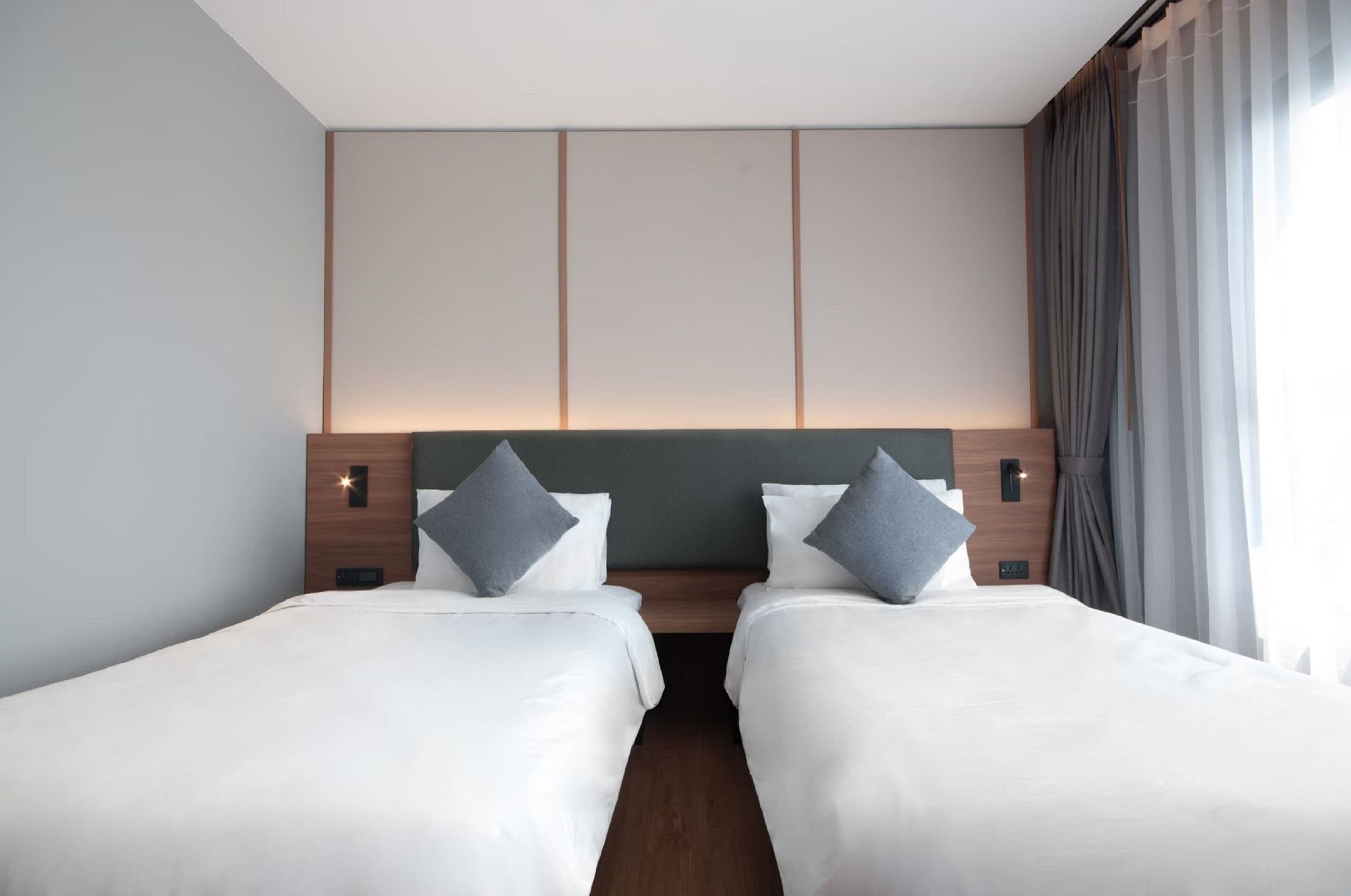 A hotel room with two single beds, a wooden headboard, a nightstand, and a window with curtains.