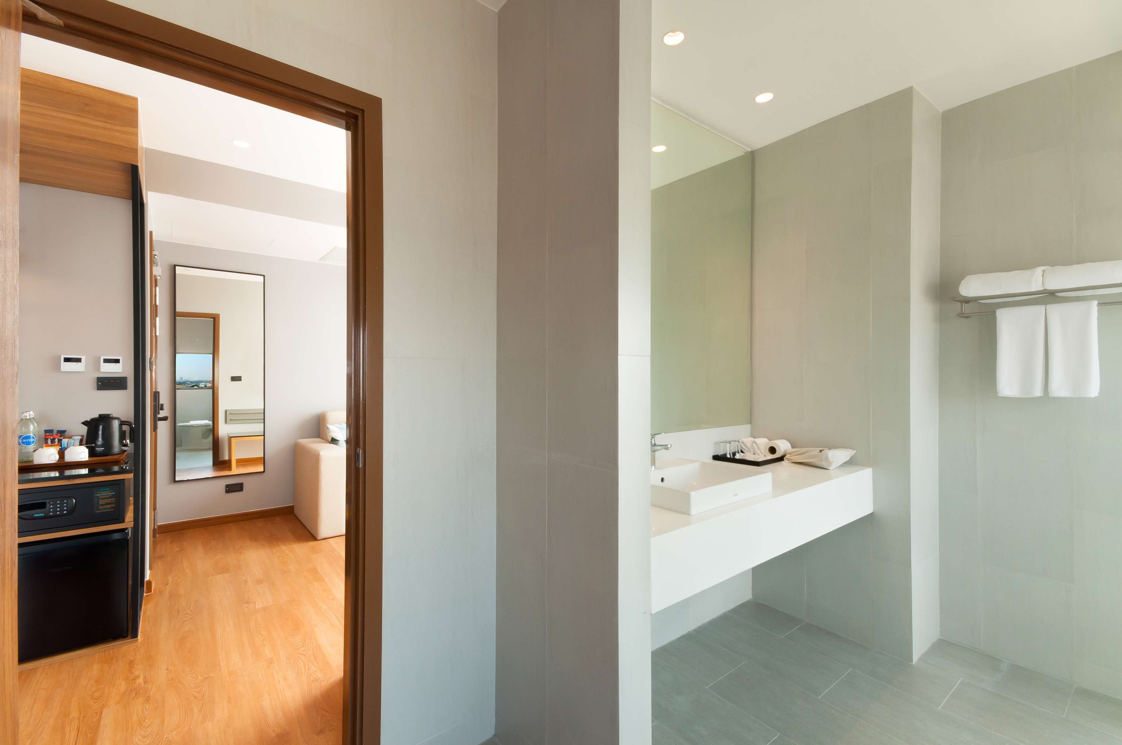 A modern hotel room with a wooden floor, a kitchenette, a large mirror, and a bathroom with a white sink and towel rack.