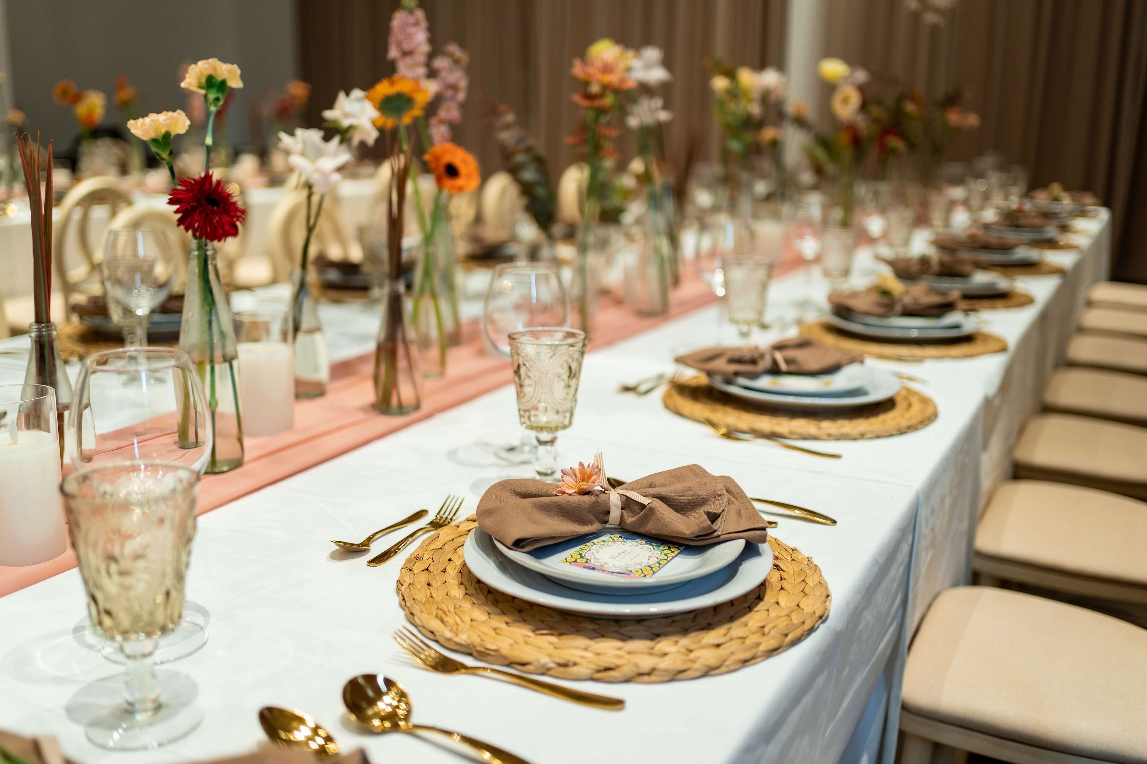 A long table set with white tablecloths, gold cutlery, and woven placemats.