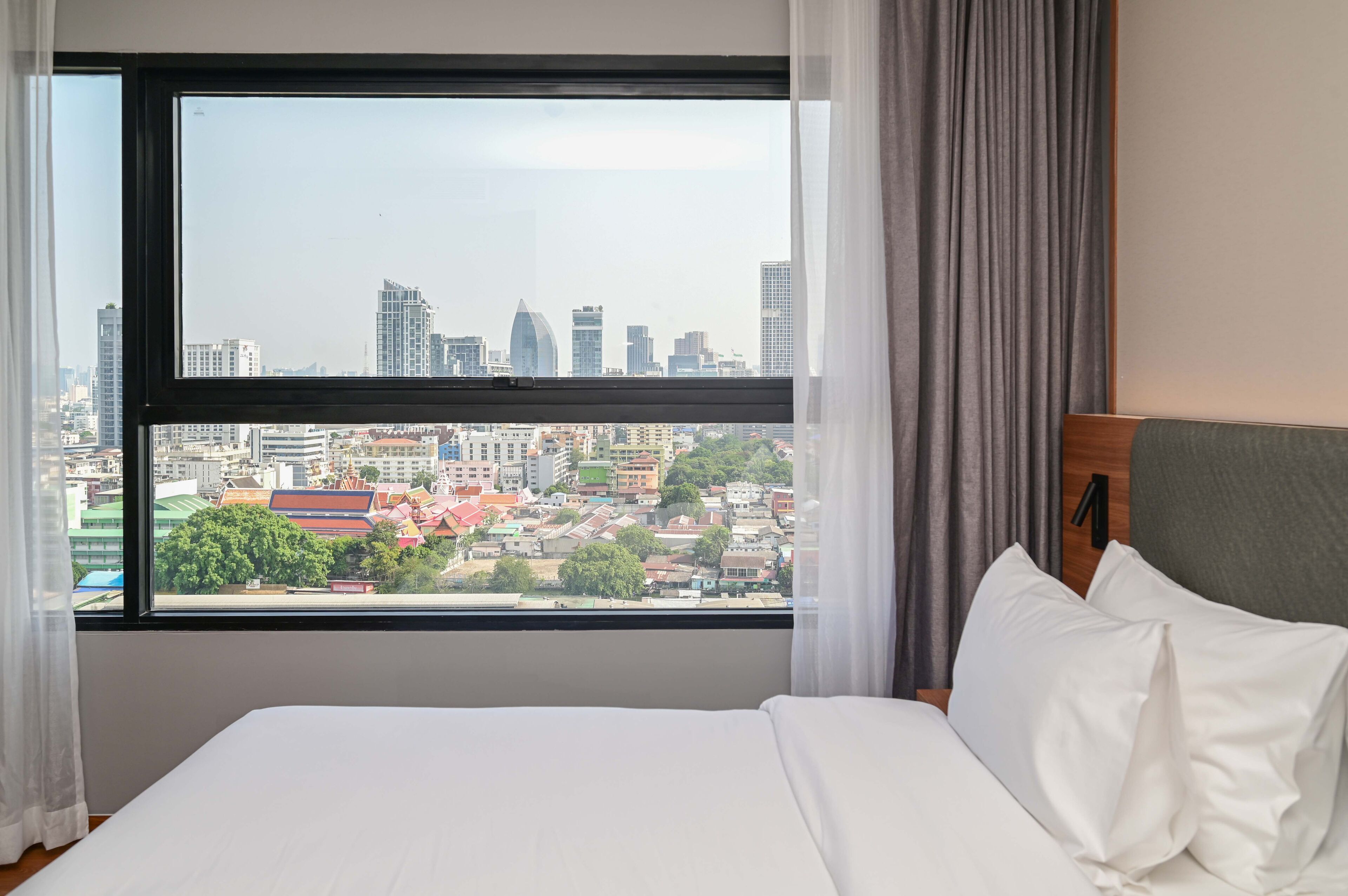 A hotel room with a large window offering a city view, a neatly made bed, and a wooden headboard.