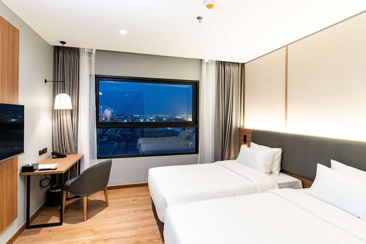 A hotel room with two beds, a desk, a chair, a TV, and a large window with a city view.