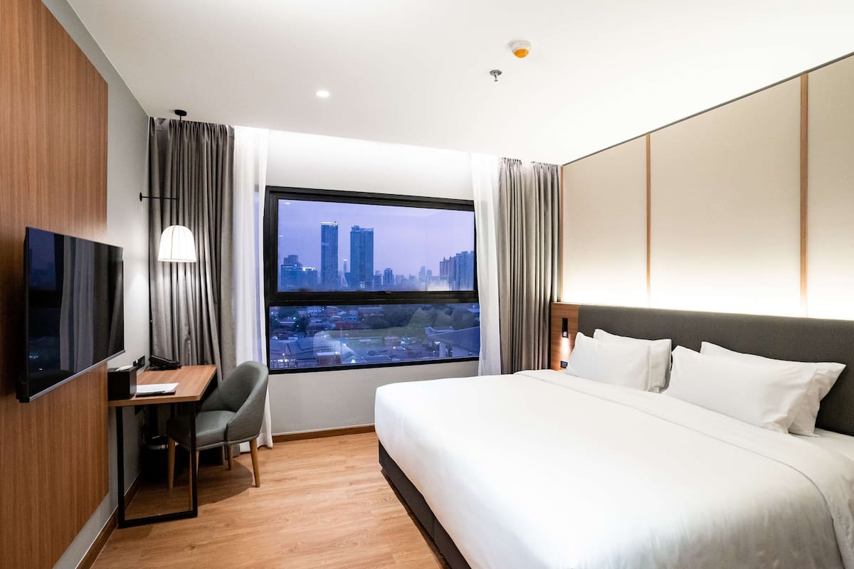 A hotel room with a large bed, a desk with a chair, a TV, and a window with a city view.