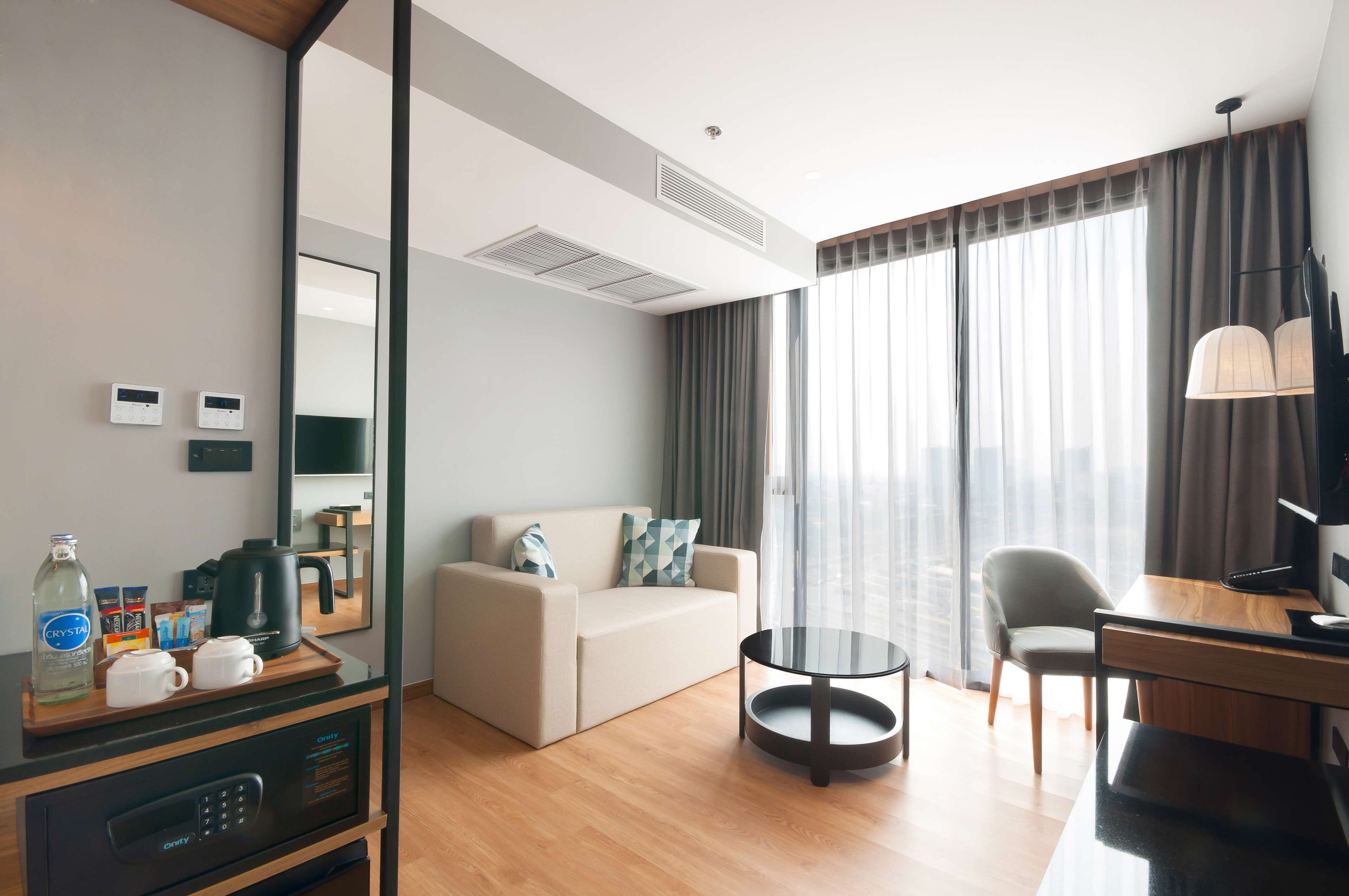 A modern hotel room with a sofa, a small round table, a desk, a mirror, and a minibar.