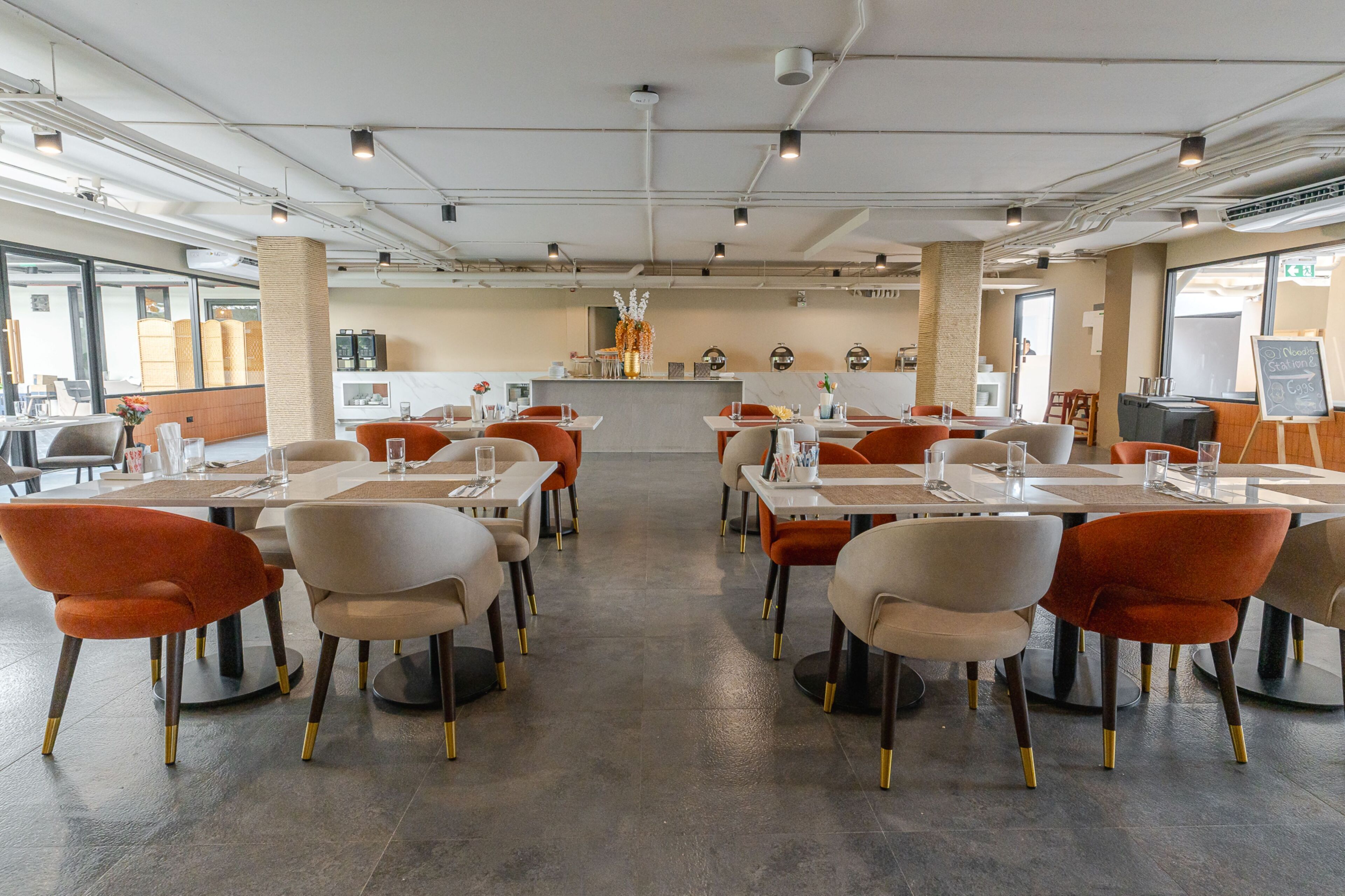 A modern restaurant with a central bar, multiple tables with orange chairs, and a view of the city through large windows.