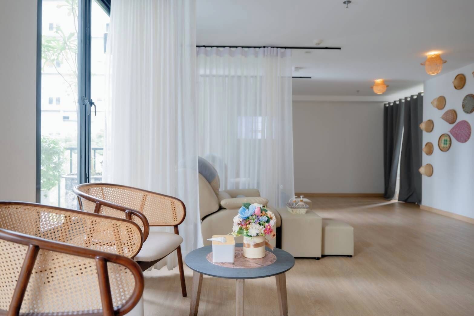 Una sala de estar moderna con un sofá, dos sillones de mimbre, una pequeña mesa redonda con un objeto decorativo y un gran ventanal con cortinas translúcidas.