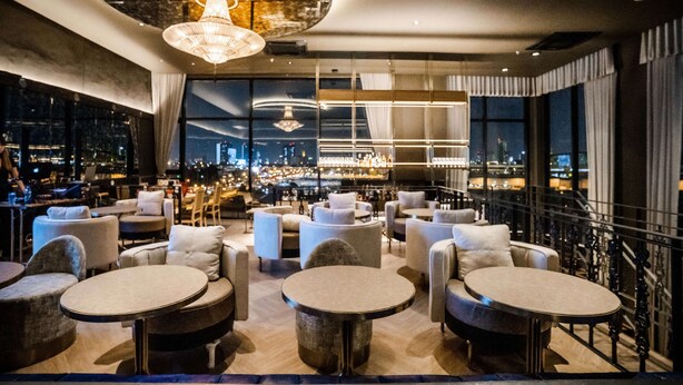 Et moderne bar- og loungeområde med runde borde og sofaer, store vinduer med udsigt over byen, samt en lysekrone.