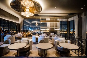 A modern bar/lounge area with round tables and sofas, large windows offering city views, and a chandelier. - Best Western Chatuchak (Bangkok)