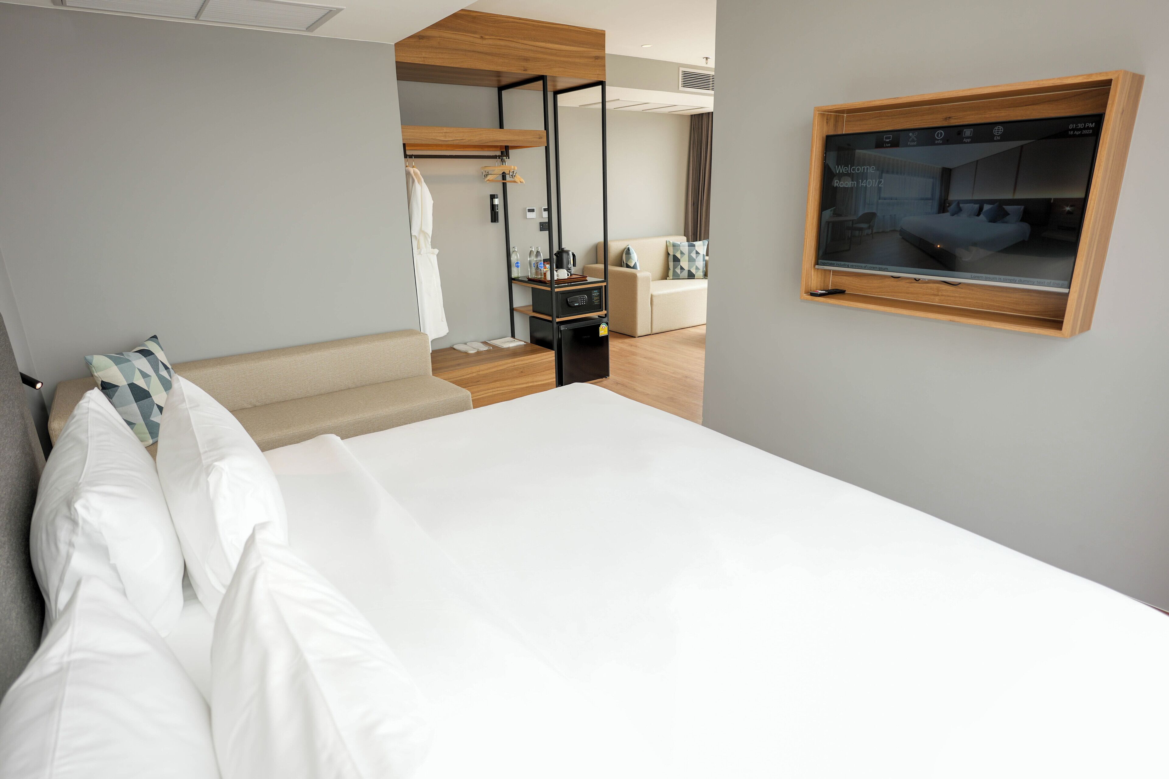 A modern hotel room with a large bed, a wooden wardrobe, a seating area, and a TV screen displaying a welcome message.