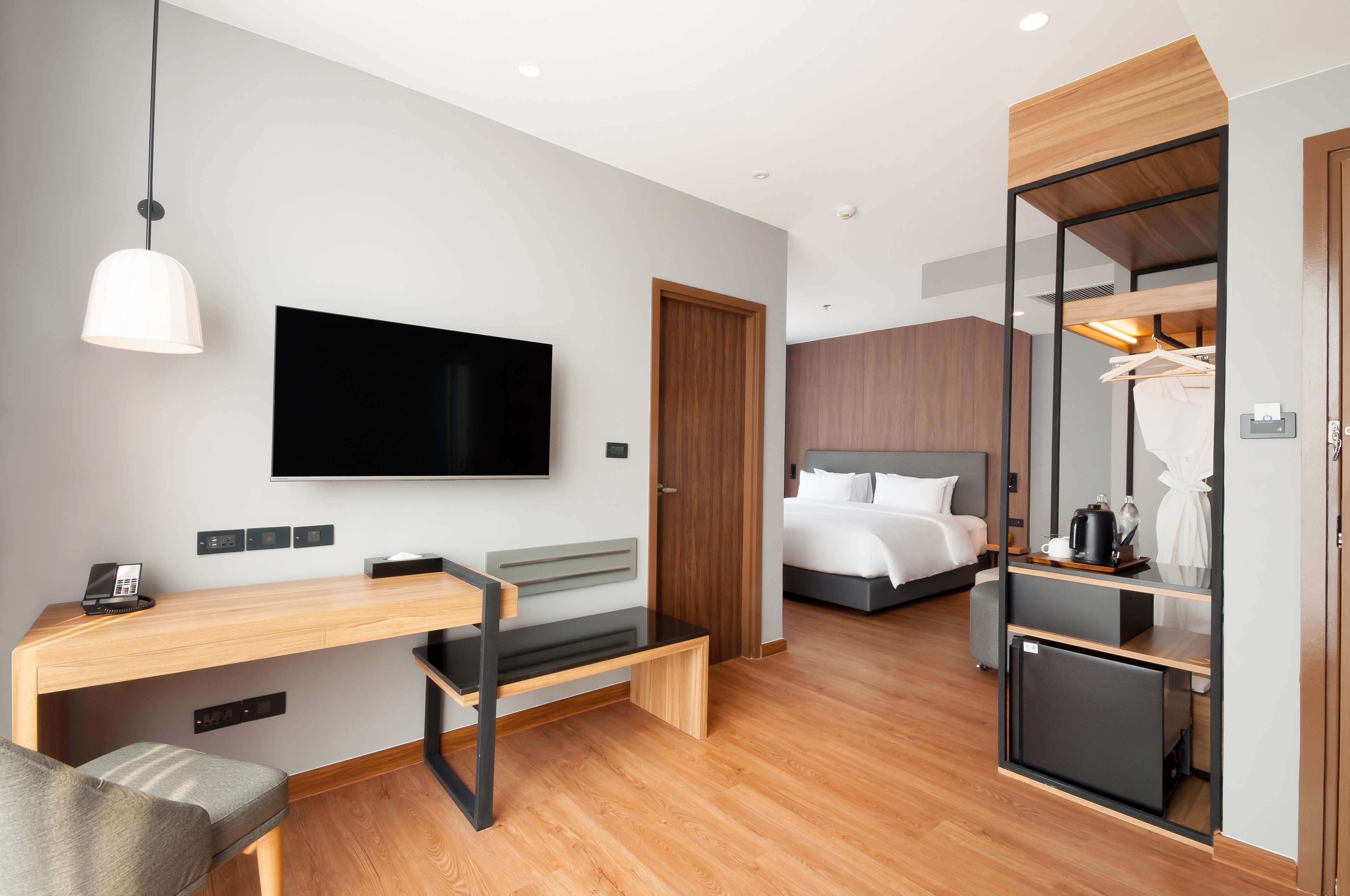 A modern hotel room with a wooden desk, a flat-screen TV, a bed with white bedding, and a walk-in closet.