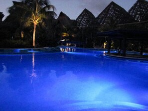 A nighttime swimming pool with a palm tree and pyramid-shaped structures in the background.