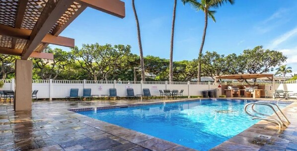 A pool area with a clear blue pool, lounge chairs, and palm trees.