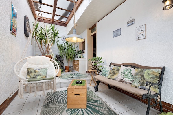 A cozy indoor space with a wooden bench, a cushioned chair, a small wooden table, and hanging plants.