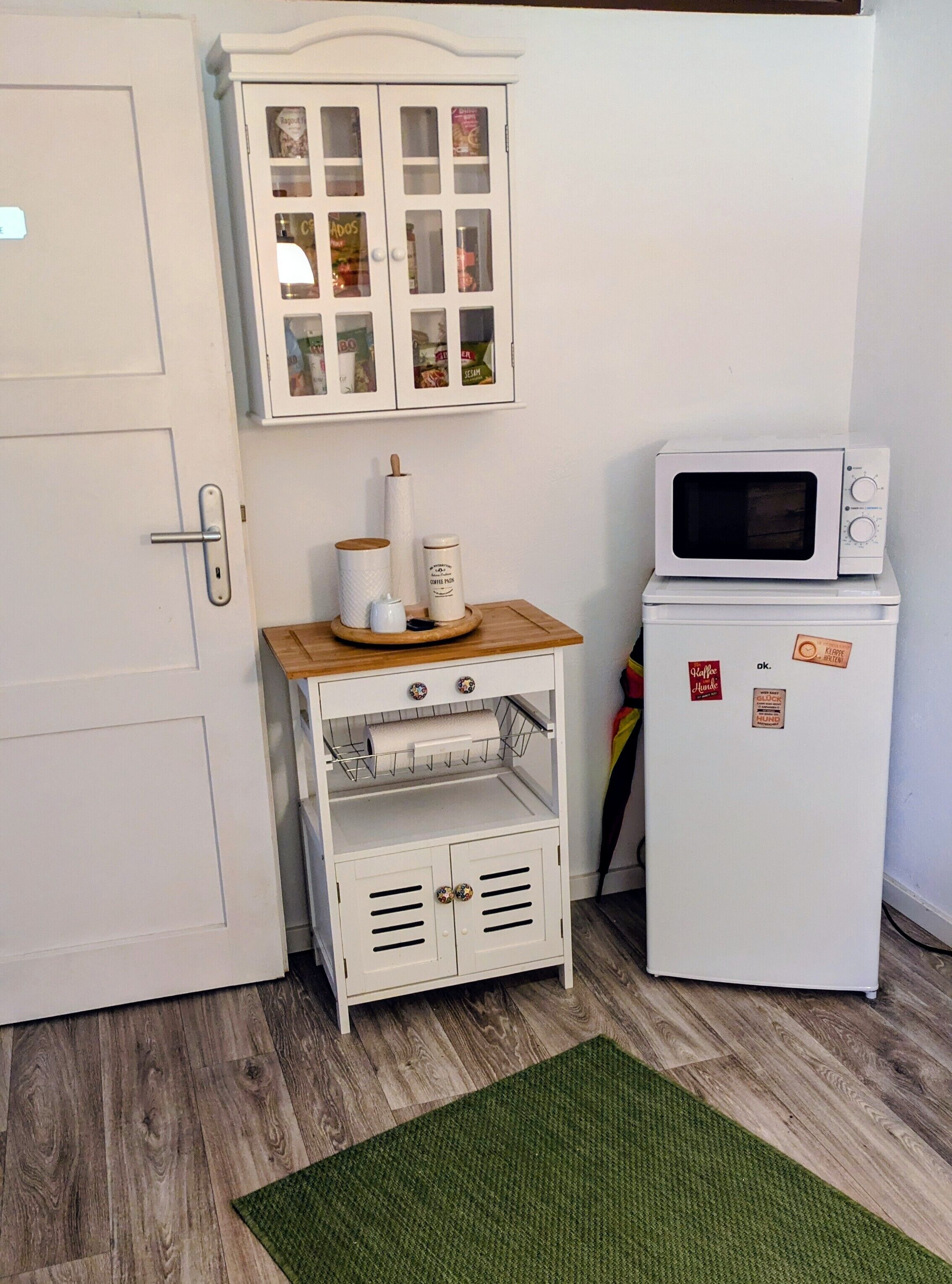 Een compacte kitchenette met een magnetron, een klein kastje met glazen deuren en een houten plank met keukengerei.