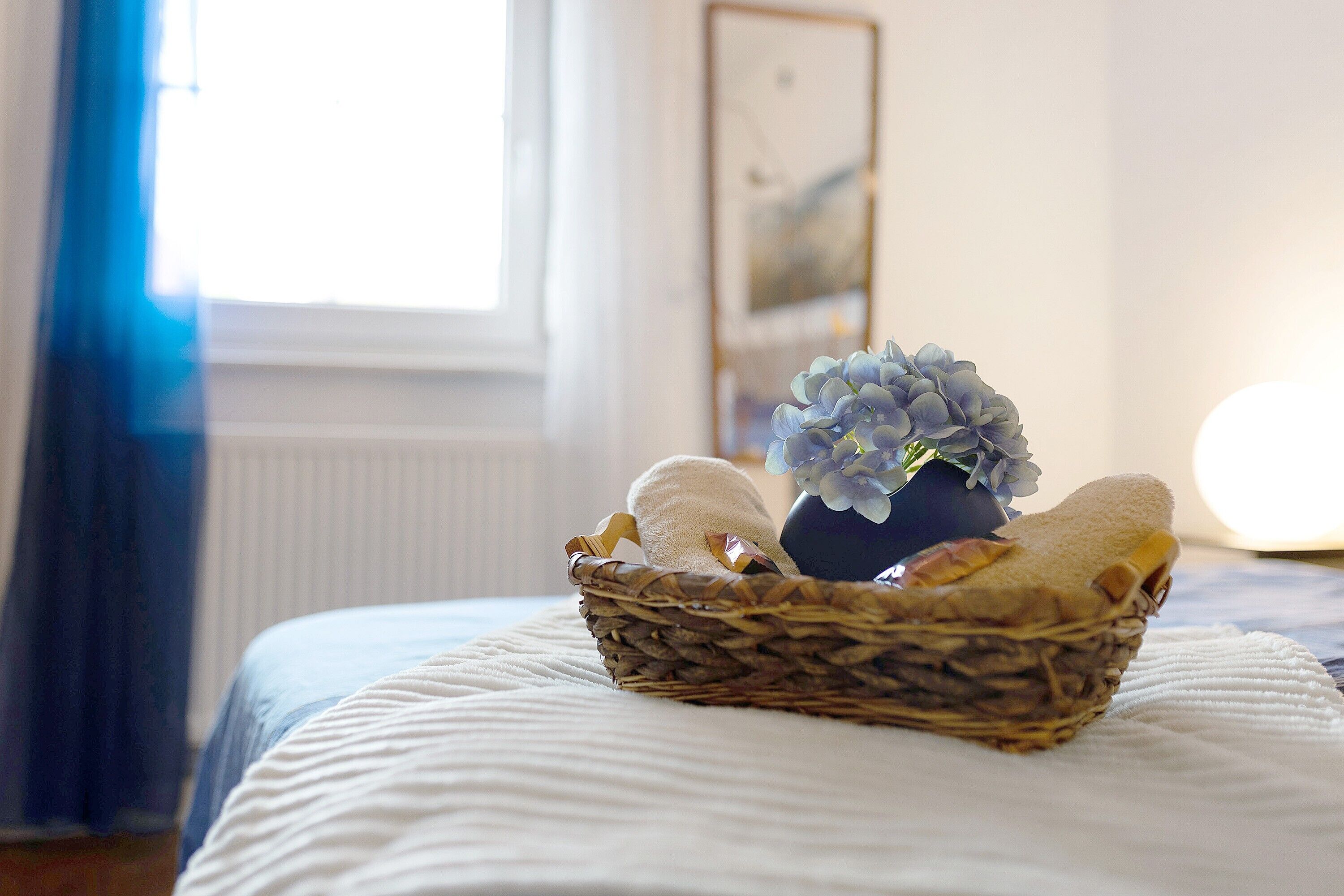 A bed with a white comforter, a wicker basket with towels and a vase of flowers on it.