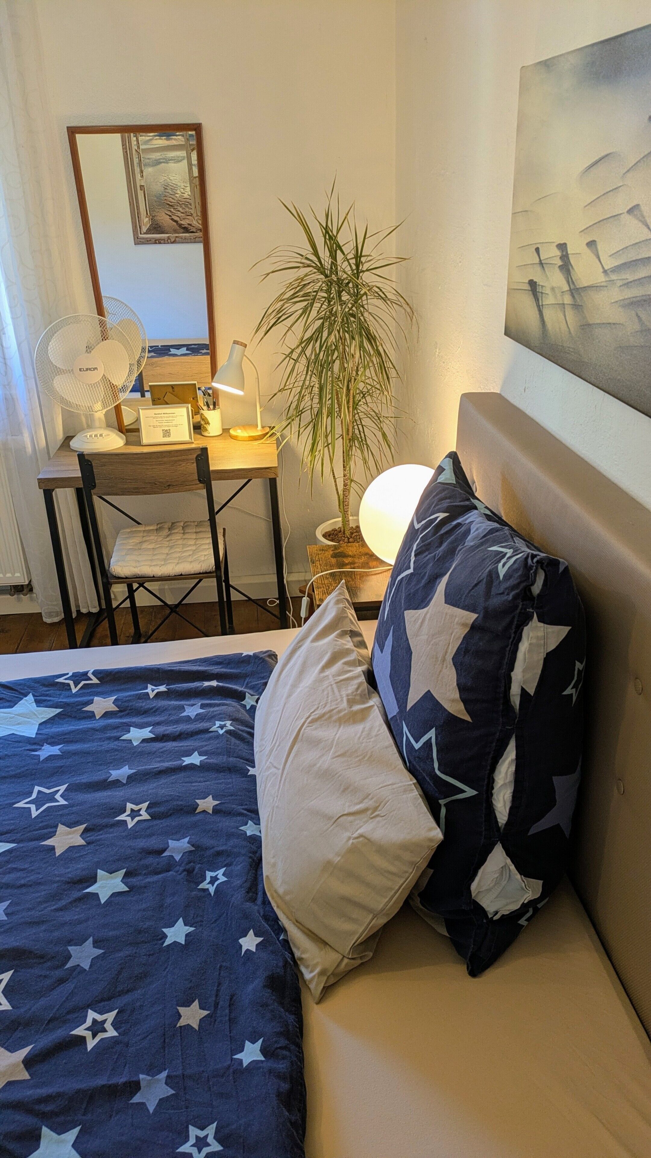 Ett sovrum med en säng, ett skrivbord, en stol, en lampa, en växt och en tavla på väggen.