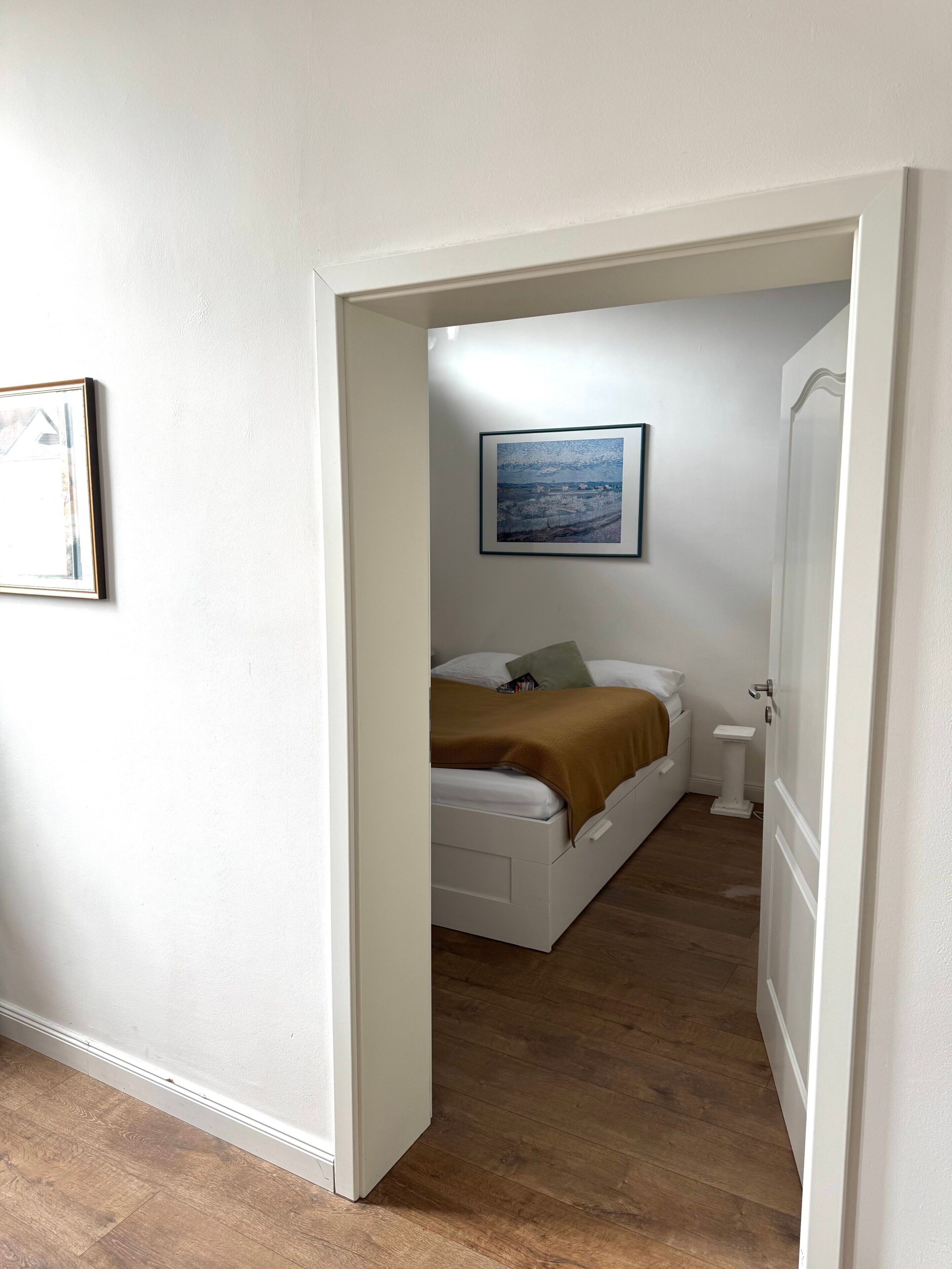 A bedroom with a bed, a bedside table, a wall-mounted picture, and a door leading to another room.