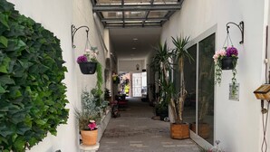Un pasillo estrecho con un banco, plantas en macetas, cestas de flores colgantes y una puerta de cristal que da a un coche.
