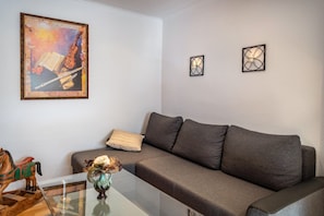 A living room with a grey sofa, a glass coffee table, a framed painting, and two wall-mounted light fixtures.