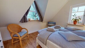 Un dormitorio con techo inclinado, una cama con almohadas y toallas dobladas, una silla de mimbre, una ventana con cortinas y una planta en maceta.