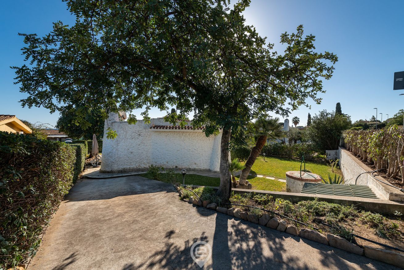 A pathway leading to a white building with a tiled roof, surrounded by greenery and a clear sky.