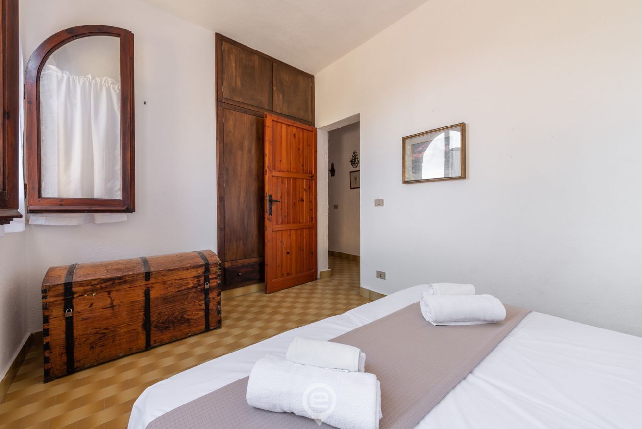 A bedroom with a bed, a wooden chest, a wardrobe, a mirror, and a framed picture on the wall.