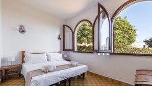 A bedroom with a large bed, a wooden bench, a nightstand, and a window with a view of greenery. - Palm Villa - Seaside Vacation (Muravera)