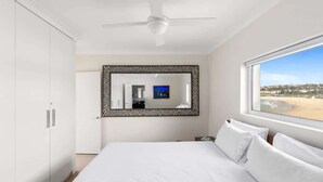 Een slaapkamer met een bed, een plafondventilator, een grote spiegel en een raam met uitzicht op het strand.