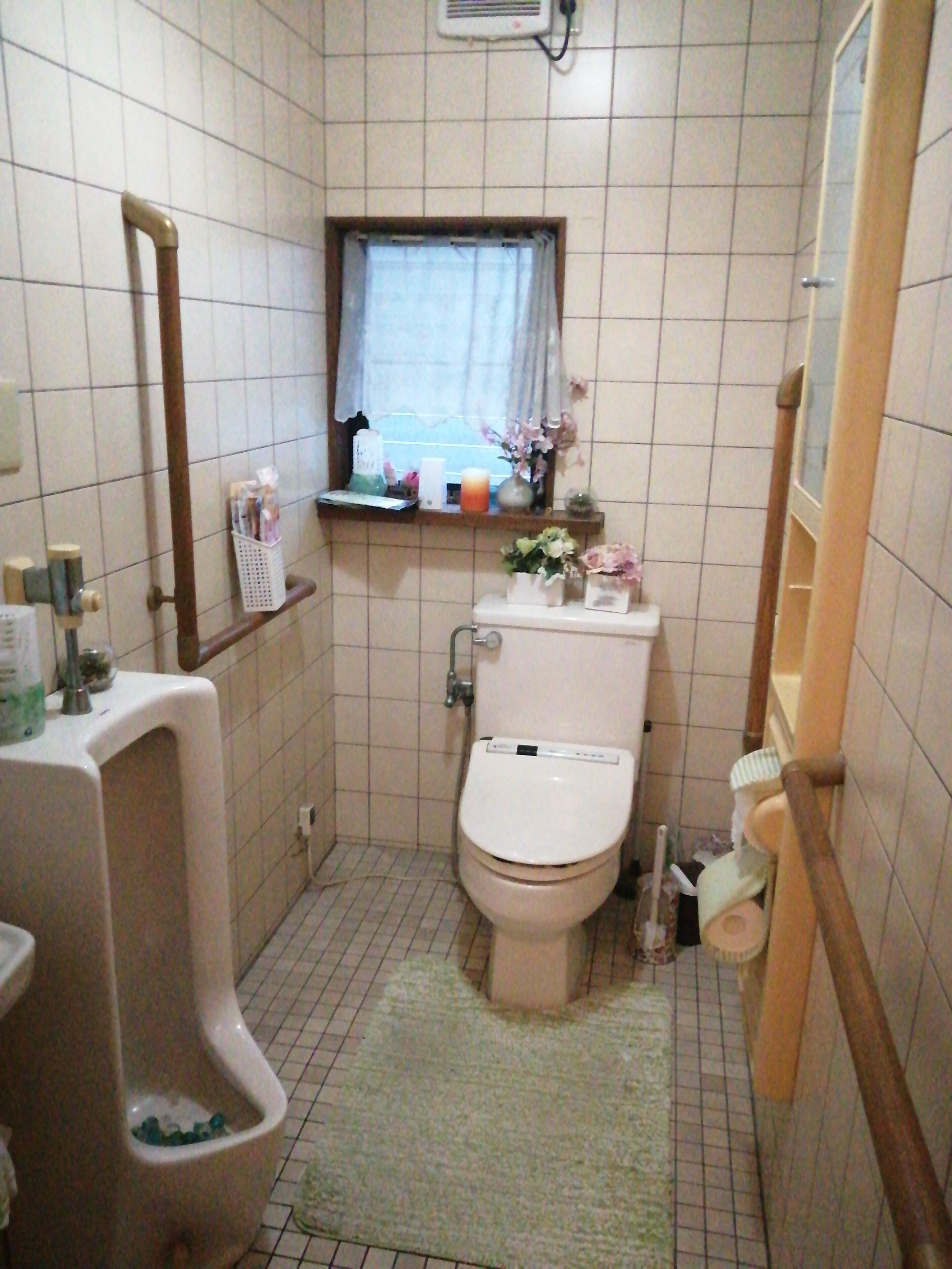 Un baño pequeño con inodoro, un orinal, una ventana con cortina y un estante con artículos de aseo.