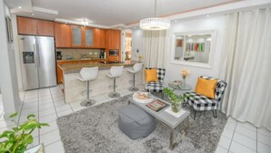 A modern kitchen with a refrigerator, microwave, and wooden cabinets. There is a dining area with a table and chairs, and a living room with a sofa and ottoman. - Tropical Haven with private pool (Carolina)