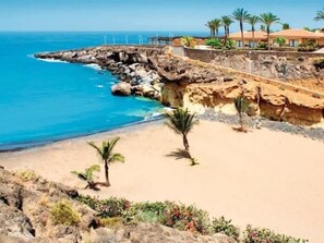 Un hotel a la costa amb una platja de sorra, penya-segades rocoses i palmeres.