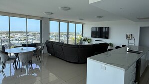 A modern living room with a sofa, dining table, and chairs. There is a large window offering a city view. - Harbour Quays Apartments (Biggera Waters)