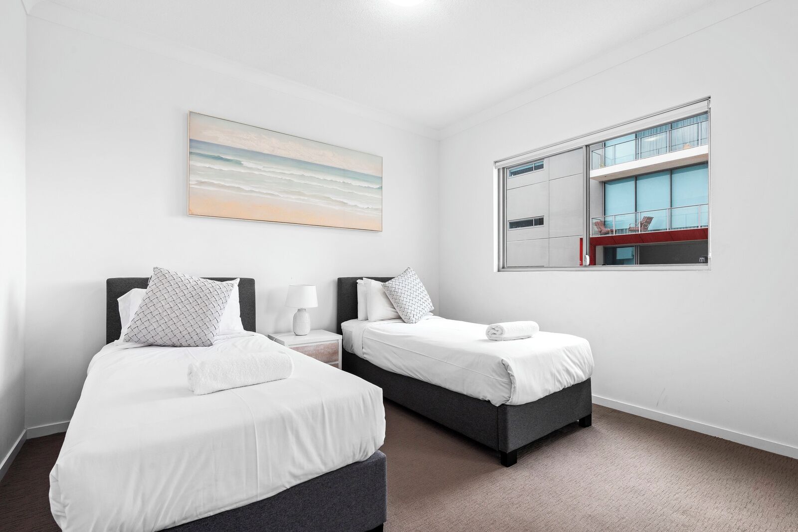 Une chambre d’hôtel avec deux lits, une grande fenêtre donnant sur un immeuble et un tableau représentant une scène de plage accroché au mur.
