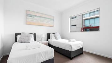 Une chambre d’hôtel avec deux lits, une grande fenêtre donnant sur un immeuble et un tableau représentant une scène de plage accroché au mur.